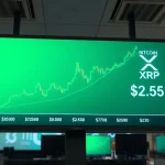Digital display showing Bitcoin and XRP price charts with green upward trend on a trading floor.
